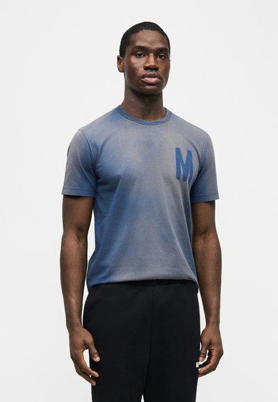 Άνδρας που φοράει ξεθωριασμένο μπλε μπλουζάκι με σκούρο μπλε "M" στο στήθος και μαύρο παντελόνι, στέκεται μπροστά σε απλό λευκό φόντο.