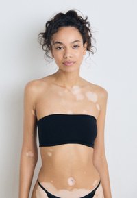 Haut noir sans bretelles avec une texture lisse, associé à un bas de bikini assorti. Le modèle a un teint moyen avec des taches claires.