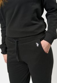 Svart bomullströja och byxor, som har en slim fit, elastisk midje och en liten vit logotyp på fickan. Slät textur.