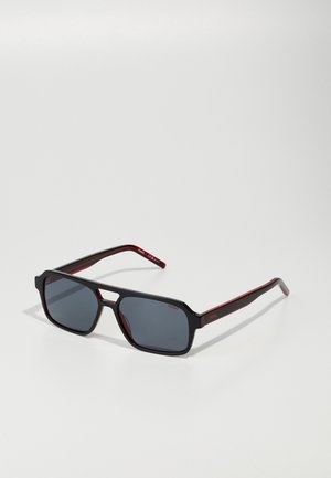 Occhiali da sole con montatura rettangolare nera e lenti grigie. Accenti rossi sulle tempie con logo del marchio. Texture liscia, design moderno.