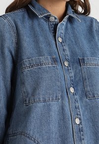 Kvinna iförd en blå jeansjacka med två bröstfickor och metallknappar fram.