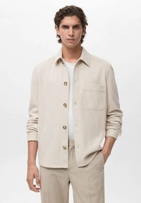 Chemise beige à boutonner avec poche poitrine, fabriquée en matériau doux. Portée par-dessus un T-shirt blanc, associée à un pantalon de couleur claire.