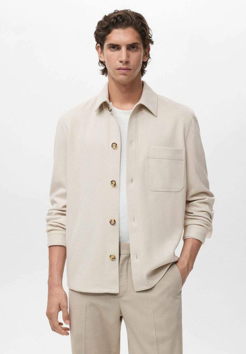 Chemise beige à boutonner avec poche poitrine, fabriquée en matériau doux. Portée par-dessus un T-shirt blanc, associée à un pantalon de couleur claire.