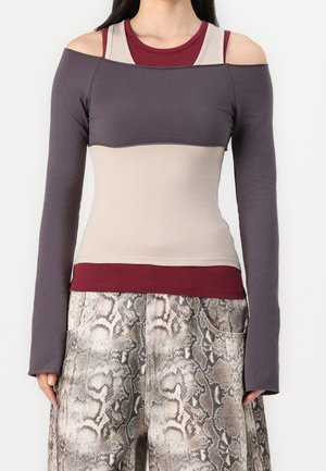 Vrouw draagt gelaagde grijze lange mouwen die van de schouder af vallen over beige en bordeauxrode bovenkleding, gecombineerd met wijde high-waist broek met slangendessin.