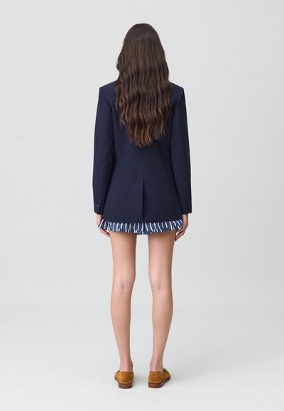 Blazer marine avec une silhouette ajustée, doté d'une fente au dos. Associé à une jupe rayée marine et blanche et des chaussures en daim marron.