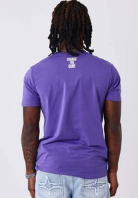 Camiseta de algodón morada con mangas cortas y cuello redondo. Texto "CAMBIA EL JUEGO" en blanco impreso en la parte superior de la espalda. Ajuste casual.