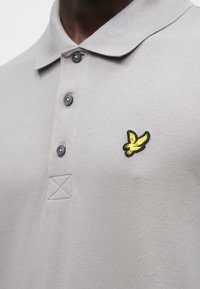 Sivá polokošeľa s textúrovanou látkou, tromi tmavými gombíkmi a žltým a čiernym vyšívaným logom vtáka na ľavej strane hrudníka.
