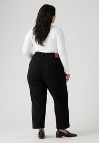 Black denim jeans with a straight leg cut, high waist, and two back pockets; paired with a fitted white long-sleeve top and black shoes.