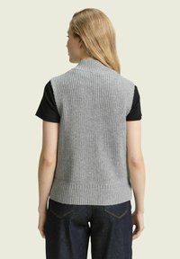 Grau gestrickte Weste mit hohem Kragen, geripptem Saum und strukturiertem Design. Über einem schwarzen Hemd getragen, hat sie eine figurbetonte Silhouette.