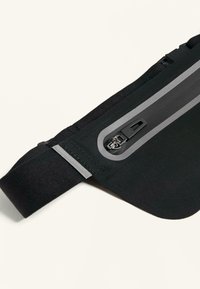 Fekete deréktáska texturált felülettel, szürke cipzárral és biztonságos csatos záródással. Állítható elasztikus pántokkal a kényelmes illeszkedésért.