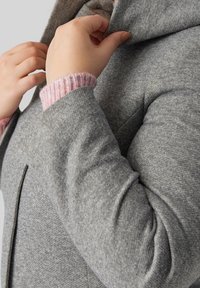 Grå uldblandet frakke med en struktureret krave, tekstureret stof og lyserøde ribstrikkede manchetter. Modellen justerer kraven og fremhæver detaljerede syninger.