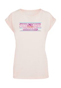 Kerge roosa puuvillane T-särk, mille varrukad on kokku rullitud, ja mille esiosas on julge "Stranger Things" logo roosa ja sinisega.
