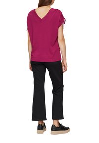 Haut fuchsia à col en V avec des manches courtes et des accents de nœud, associé à un pantalon noir court et des baskets noires épaisses sur un fond uni.