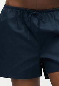 Σκούρα μπλε σορτς με κορδόνι στη μέση, που φοριούνται από άτομο με σκουρόχρωμο δέρμα, με ελαστική μέση και πλευρική σχισμή στο τελείωμα.