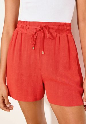 Shorts rouges mi-longue taille élastiquée froncée et cordon noué, portés avec un haut blanc, montrés de la taille jusqu'au milieu de la cuisse.