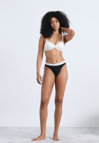 Reggiseno bianco con design sagomato e spalline sottili, abbinato a slip bikini neri con fascia in vita con logo bianco. Tessuto liscio ed elastico.