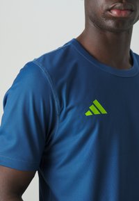 Blue athletic shirt made of smooth fabric with green Adidas logo, crew neck design, and short sleeves, showcasing detailed stitching.