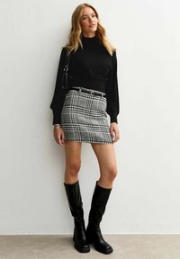 Donna che indossa un top nero a maniche lunghe, una mini gonna a quadri bianco e nero con cintura, stivali neri al ginocchio, in posa contro un muro bianco.