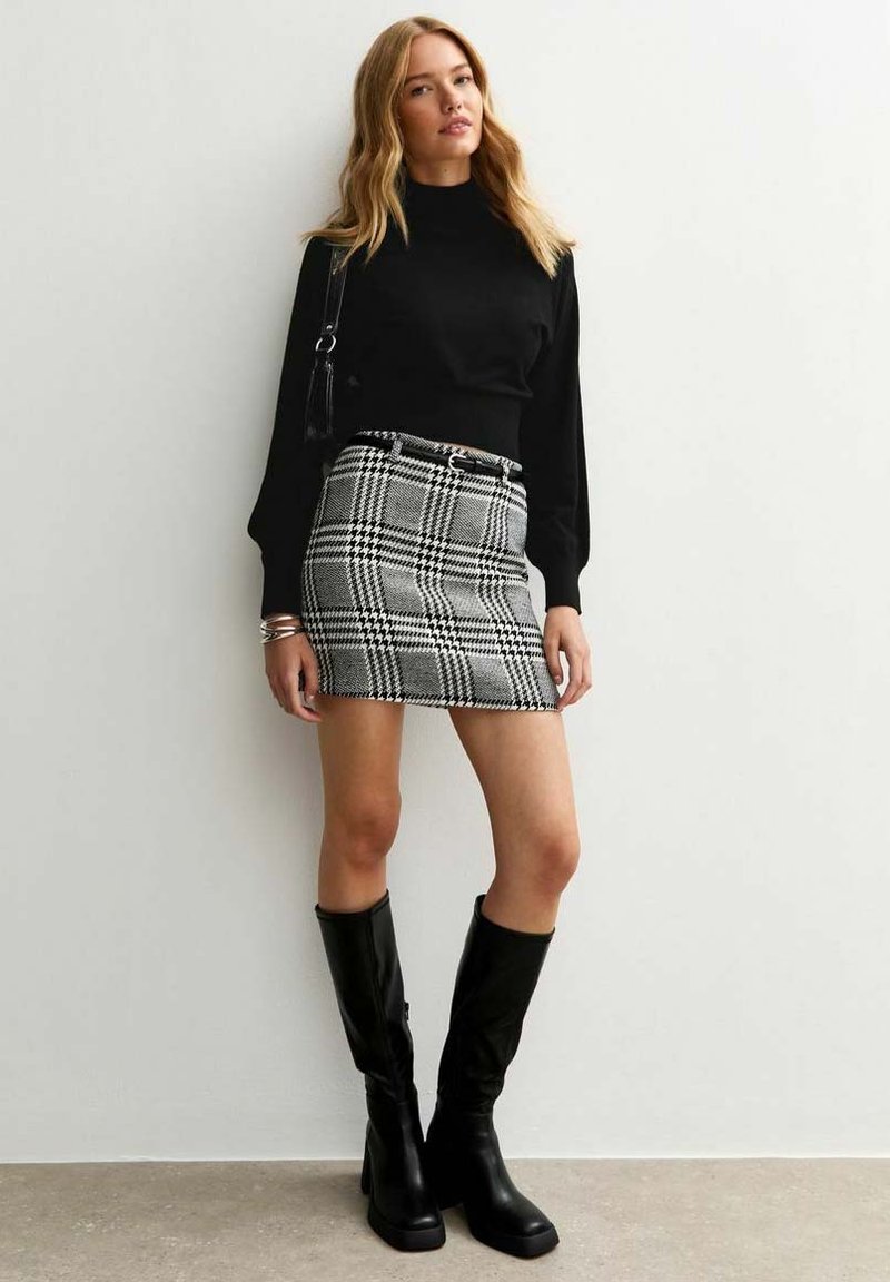 Donna che indossa un top nero a maniche lunghe, una mini gonna a quadri bianco e nero con cintura, stivali neri al ginocchio, in posa contro un muro bianco.