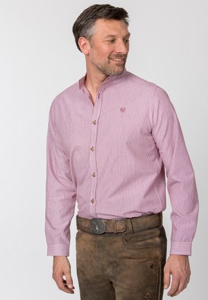 Mann trägt rosa gestreiftes Hemd mit Knöpfen und braune Lederhose mit dekorativer Gürtelschnalle, blickt nach links und lächelt leicht.