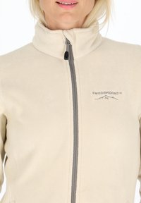 Beige fleece jasje met een hoge kraag, grijze rits en geborduurd logo. Zachte textuur, aansluitend design en twee zijzakken.