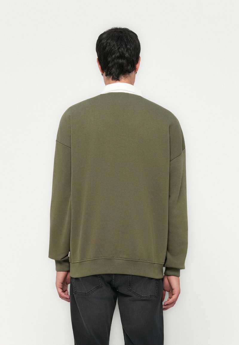 Olivgrüner Sweater mit lockerer Passform, gerippten Bündchen und Ausschnitt; hat ein Etikett an der unteren Seite; kombiniert mit grauen Jeans.