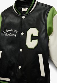Chaqueta varsity de imitación de cuero negra con mangas verdes y blancas. Presenta bordado de "Champs Academy" y una letra "C" en aplicación. Diseño de cierre delantero con botones.