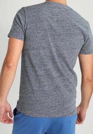 Man met een eenvoudig grijs T-shirt met korte mouwen en blauwe broek, van achteren gezien tegen een effen achtergrond.