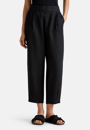 Pantalones de lino negros con cintura alta, que presentan pliegues y aberturas de piernas ajustadas. Combinados con sandalias de dedo texturizadas negras.