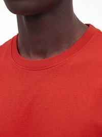Nærbillede af en persons nakke og underansigt iført en ensfarvet rød t-shirt med rund hals mod en hvid baggrund.