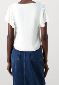 Vit, kortärmad topp med fyrkantig halsringning och mjukt volangärm. Avstämmer med en denimkjol som har bakfickor.