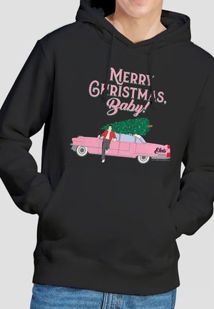 Person mit schwarzem Kapuzenpullover neben pinkem Auto, Mann lehnt an Auto und trägt Weihnachtsbaum, dazu der Text "Frohe Weihnachten, Baby!"