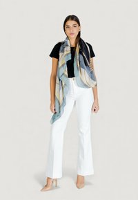 Sciarpa di seta in tonalità blu e beige con motivi geometrici, drappeggiata intorno al collo di una modella che indossa una maglia nera e pantaloni bianchi a zampa d'elefante.