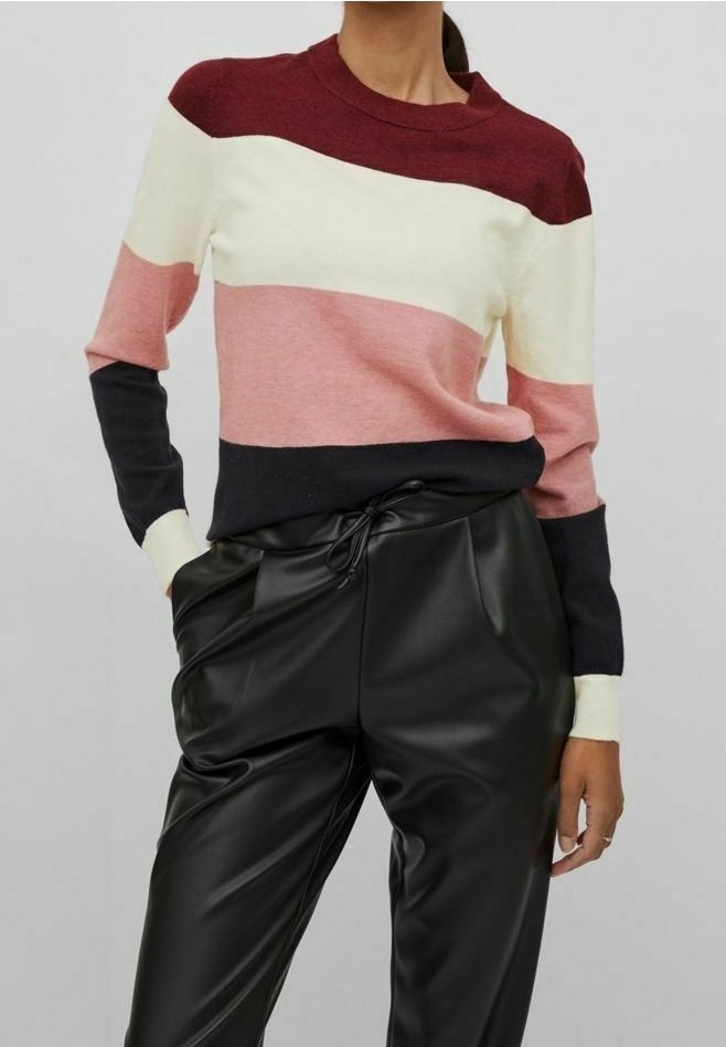 Femme portant un pull à manches longues bicolore en bordeaux, crème, rose et noir, associé à un pantalon noir en similicuir avec taille à cordon.