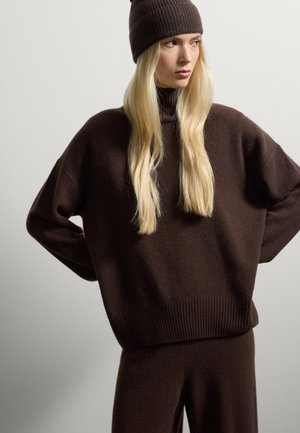 Pull en tricot marron avec un col montant et un ourlet côtelé, accompagné d'un bonnet côtelé assorti. Texture douce, coupe décontractée et manches longues.