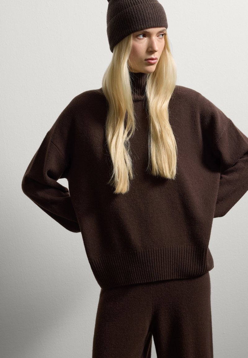 Bruine gebreide trui met een hoge hals en ribbelrand, gecombineerd met een bijpassende ribbelbeanie. Zachte textuur, ontspannen pasvorm en lange mouwen.