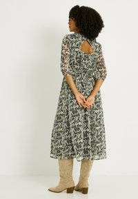 Vestido verde fluido con patrones negros, mangas tres cuartos y un detalle de abertura en la espalda, combinado con botas de cowboy beige texturizadas.