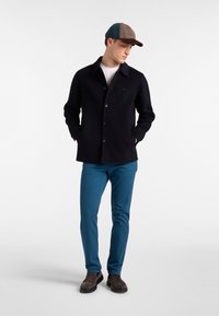 Giacca di lana blu navy con collo a camicia e chiusura a bottoni, abbinata a pantaloni teal e scarpe in camoscio marrone scuro, e un cappello a fantasia.
