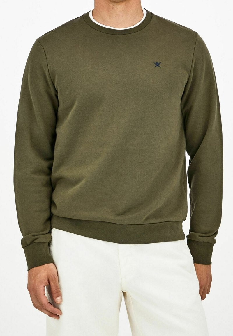 Homme portant un sweat-shirt vert olive à manches longues et un pantalon blanc, debout devant un fond blanc.