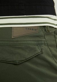 Plano detalle del bolsillo trasero de unos pantalones verdes con una etiqueta de cuero marrón con la inscripción "CHASIN'" y el dobladillo de una camisa a rayas en negro, blanco y verde encima.