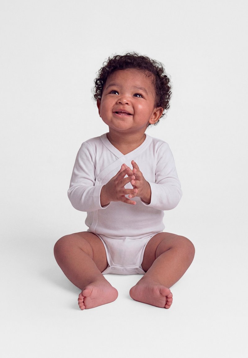 Bébé souriant aux cheveux bouclés assis par terre, portant un body blanc à manches longues, les mains jointes, regardant légèrement vers le haut.