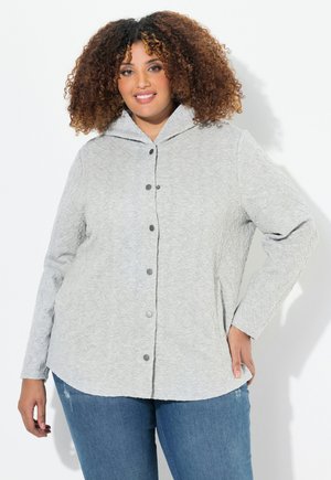 Veste en chemise matelassée grise avec capuche, dotée d'une fermeture à boutons et de manches longues, portée avec un jean bleu. Tissu texturé, coupe décontractée.