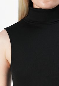 Detailní záběr osoby v bezrukávovém černém žebrovaném roláku, ukazující krk, rameno a část obličeje a paže.