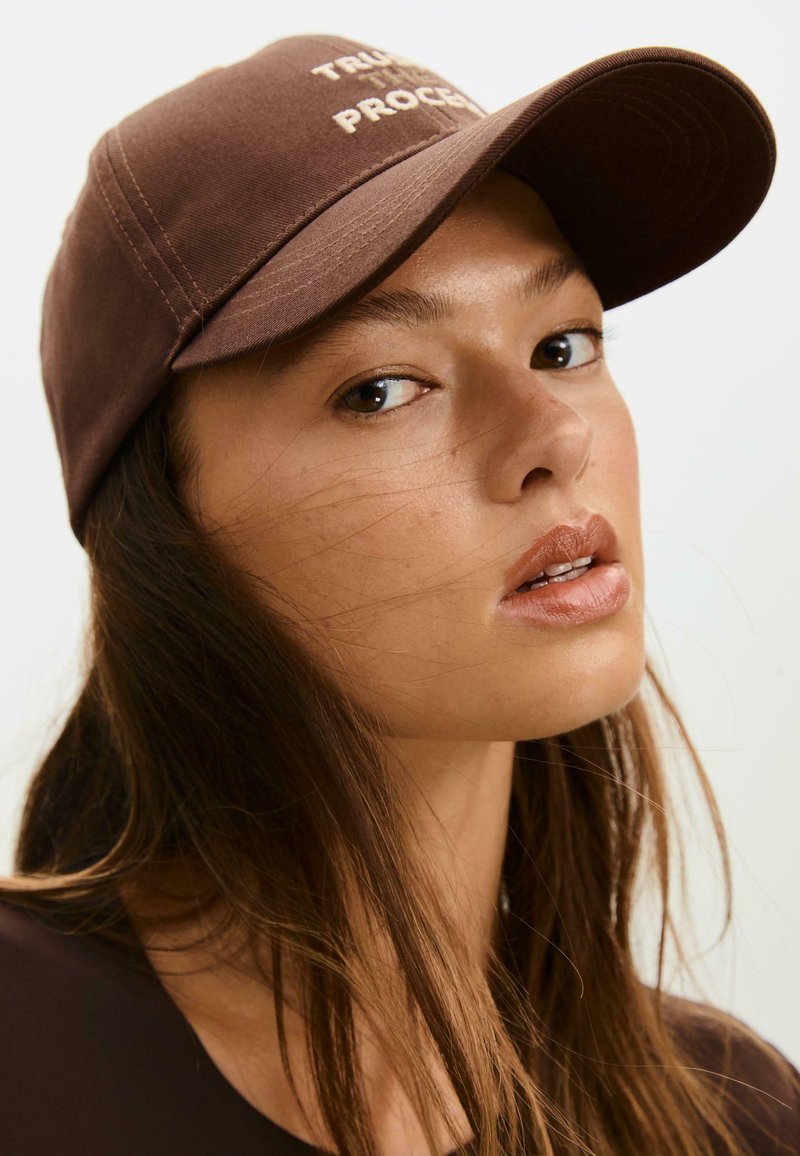 Cappellino da baseball marrone con visiera curva e testo "AFFIDATI AL PROCESSO". Presenta dettagli di cucitura sottili e una texture di tessuto liscia.
