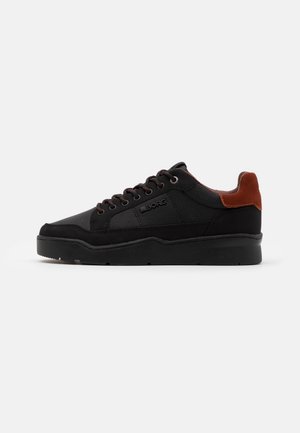 Sort low-top sneaker med sorte snørebånd, tekstureret sidelpanel og brun læderhælkappe på hvid baggrund.
