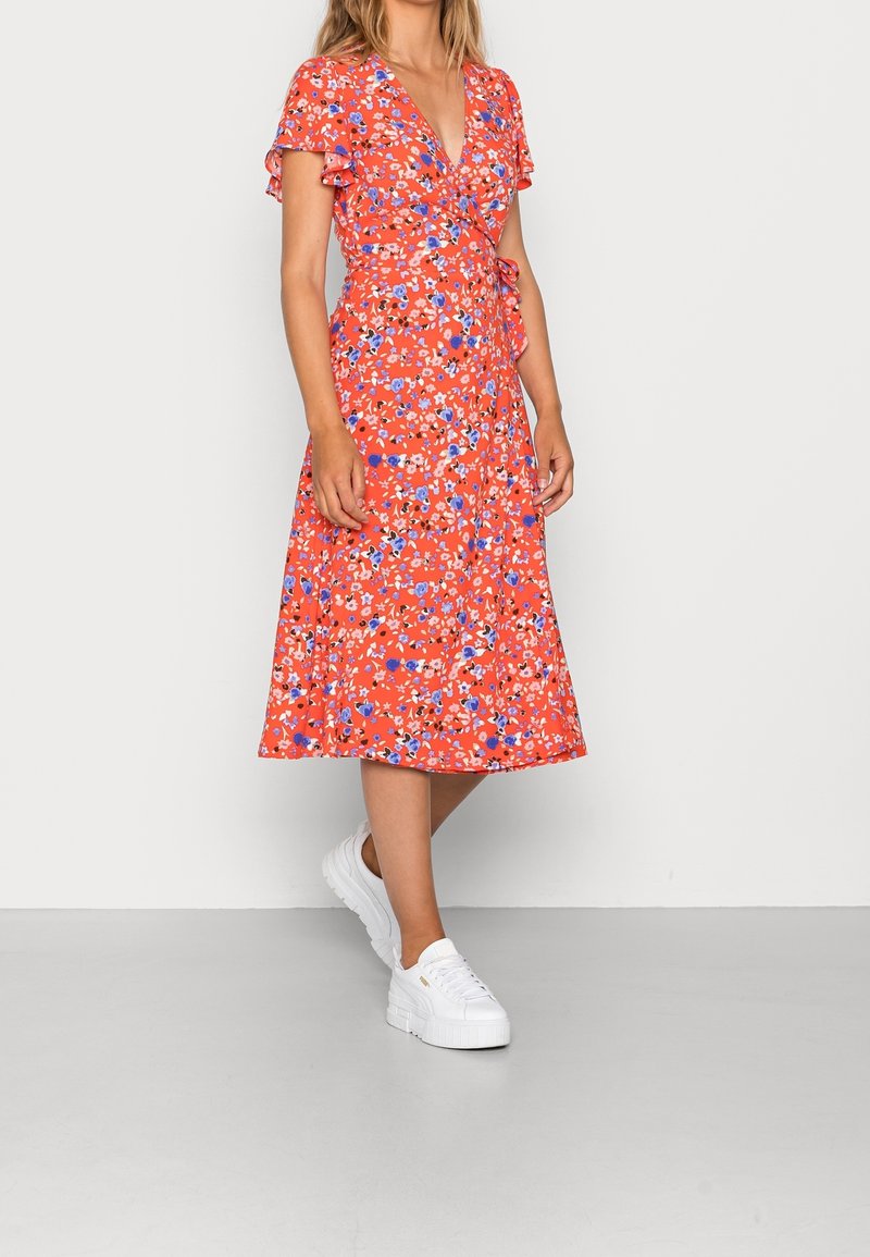 Femme portant une robe portefeuille rouge à fleurs avec des manches volantées et des baskets blanches à plateforme, debout devant un fond clair uni.