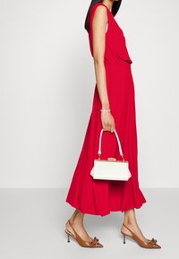 Czerwona sukienka bez rękawów z marszczeniem w talii, połączona z brązowymi sandałami na obcasie z kokardą oraz usztywnioną białą torebką z złotymi akcentami.