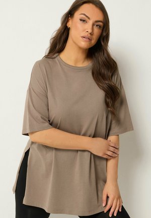 Woman with long brown hair wearing oversized taupe t-shirt, standing with one arm crossed and neutral facial expression against plain background.