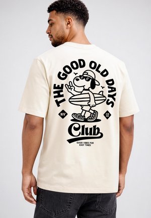 Man wearing cream T-shirt with black graphic of dog holding surfboard and text "The Good Old Days Club, good vibes for easy times."