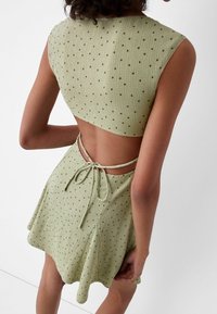Vestito verde a coste con top corto, schiena scoperta e chiusura a fiocco. Presenta piccoli punti scuri su tutta la stoffa, con design della gonna fluttuante.